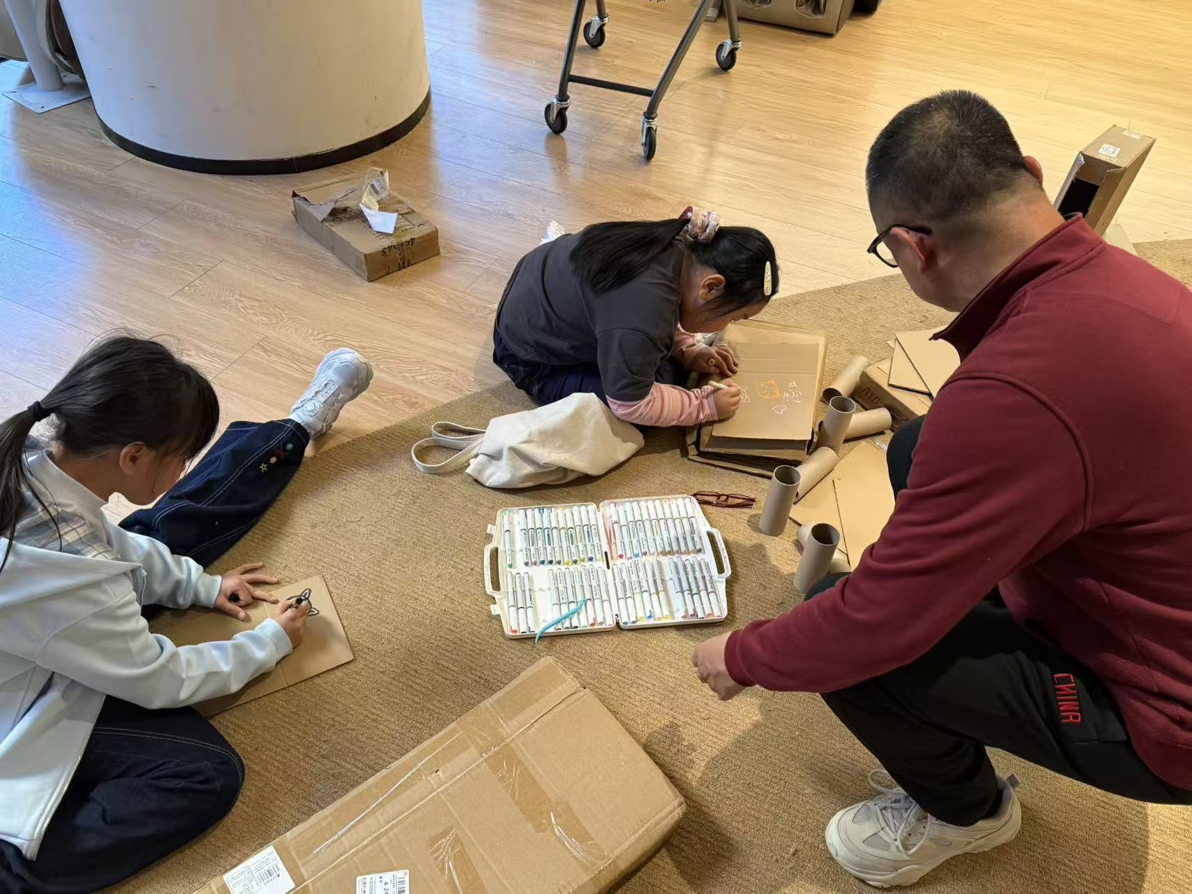 Children drawing on cardboard while another participant sits beside them.